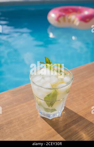 Refreshing Mojito cocktail with fresh lemon and mint leaves with ice cubes placed on wooden board near swimming pool in summer day
