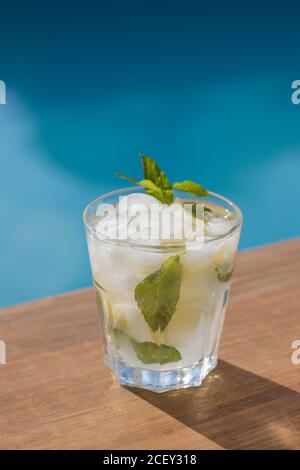 Refreshing Mojito cocktail with fresh lemon and mint leaves with ice cubes placed on wooden board near swimming pool in summer day Stock Photo