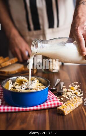 Crop anonymous housewife pouring yogurt from glass bottle into bowl with cereals and banana while preparing healthy breakfast at home kitchen Stock Photo