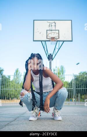 Full length of young African American female with braids wearing trendy ripped jeans and white tank top with sneakers sitting against basketball hoop on playground and looking at camera Stock Photo
