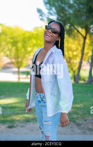 Side view of optimistic African American millennial female in sunglasses dressed in stylish white blouse over black top and trendy ripped jeans standing in green summer park Stock Photo