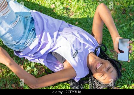 High angle of modern hipster African American female with Afro pigtails lying on green grass and browsing smartphone while resting in park in summer day Stock Photo