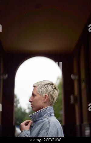 Side view of calm female model wearing denim jacket standing in arched passage on street and looking away Stock Photo