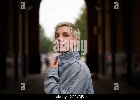 Side view of calm female model wearing denim jacket standing in arched passage on street and looking at camera Stock Photo