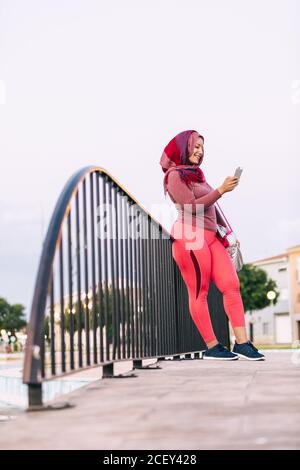 Low angle side view of Arab sportsWoman in hijab leaning on metal railing on street and chatting on social media after training Stock Photo