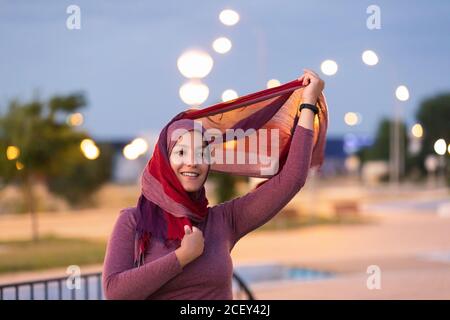 Delighted ethnic female standing on street in evening and putting on hijab while looking away Stock Photo