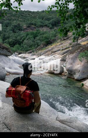 Back view of anonymous male hiker sitting on stone near rapid river and enjoying magnificent mountainous terrain Stock Photo