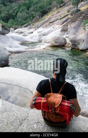 Back view of anonymous male hiker sitting on stone near rapid river and enjoying magnificent mountainous terrain Stock Photo
