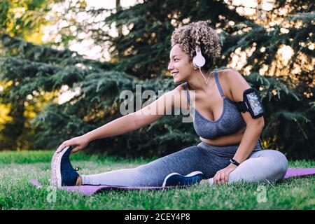 Happy young ethnic female with curly hair in sportswear listening to music with headphones and smartphone and doing seated forward bend exercise while stretching body during fitness training in summer park