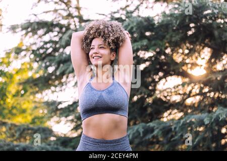 low angle of positive energetic young ethnic female with afro hairstyle wearing gray top and leggings stretching with arms behind head while standing near green trees during training in park 2cey4pm