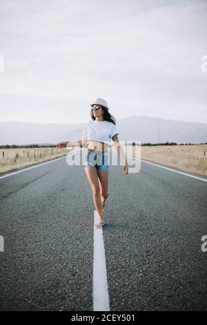 Serene female traveler in summer wear walking along empty asphalt roadway on background of mountainous landscape and enjoying freedom Stock Photo