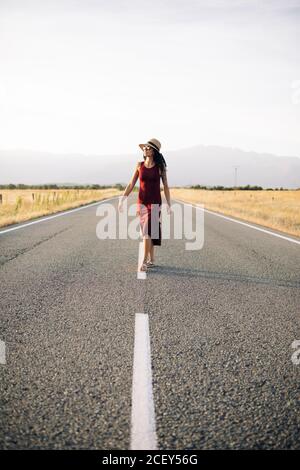 Carefree female traveler in summer dress and sunhat walking along empty roadway and looking away while enjoying freedom Stock Photo