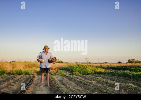 Aged male farmer standing barefoot with wicker basket of fresh fruits and vegetables on garden bed and looking away Stock Photo