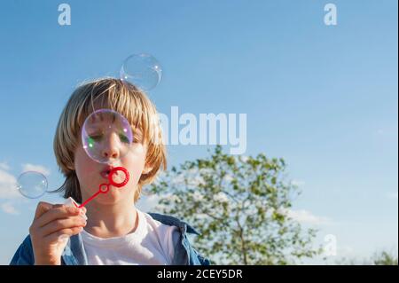 Cute child standing in park and blowing soap bubbles while entertaining during summer weekend Stock Photo
