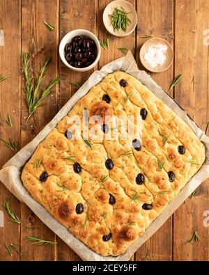 Top view of traditional Italian whole focaccia bread with olives and rosemary and salt arranged on wooden table Stock Photo