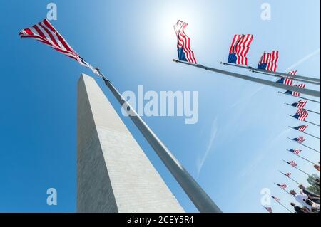 From below of Washington Monument surrounded by pillars with American flags fluttering on wind against blue cloudless sky in National Mall Stock Photo