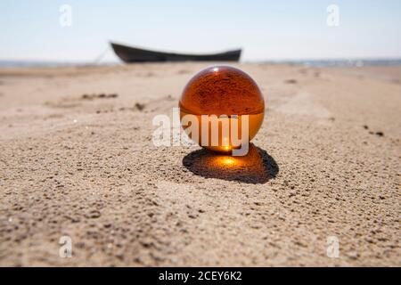 Orange glass ball on sand near the sea. Abstract conceptual photo. Stock Photo