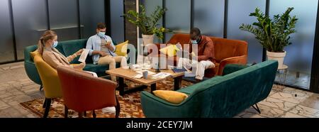 Business team during the COVID-19 outbreak, young multiracial office workers wearing medical protective masks working together while sitting in lounge Stock Photo