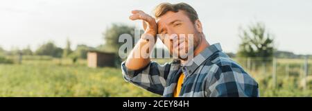 panoramic shot of exhausted farmer touching forehead while standing in field and looking at camera Stock Photo