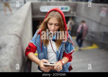 Woman in red hoodie is reading messages on the smartphone screen standing near subway enter. Young caucasian girl near underground station. Stock Photo