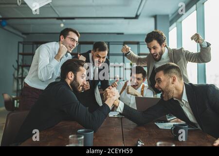 joyful coworking business men in tuxedo, cheerfully playing arm wrestling after working day in office Stock Photo
