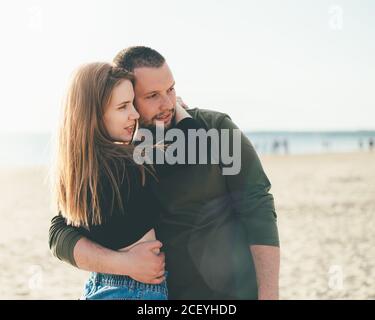 Young adult couple standing on coast and huging each other and looking away to someone. Handsome smiling man embracing beautiful smiling woman. Copy space Stock Photo