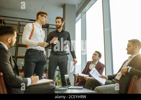 two young caucasian bearded men in formal shirts stand giving speech to co-workers, holding paper document in hands. isolated in modern boardroom, sta Stock Photo