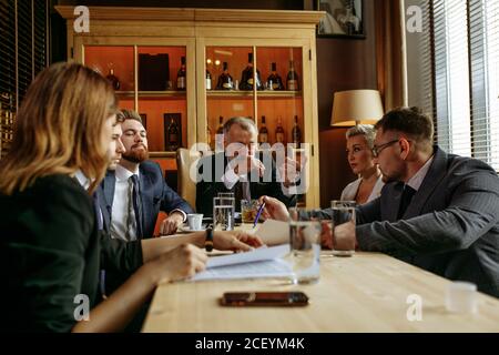 Aged boss sitting at head of wooden table in leather brown chair and pointing to young team member in glasses. Business partners watching out for lead Stock Photo