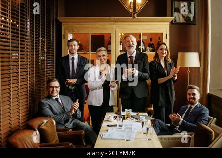 old businessman in black formal jacket and white shirt surrounded by young joyful colleagues applauding at camera with closed eyes on background of br Stock Photo