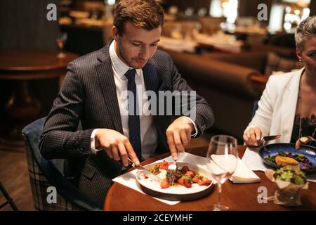 Close up of young pleasant man with stylish haircut, in formal nice suit, taking a piece of food by silver fork, sitting on soft chair at well settled Stock Photo