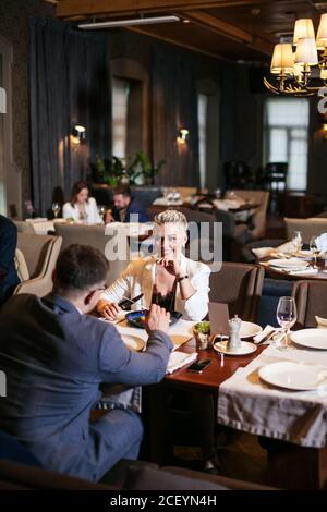 Young lovers have dinner in crowded restaurant, pleasant woman keeps chin, smiles at shy man with amorous look, vertical shot Stock Photo