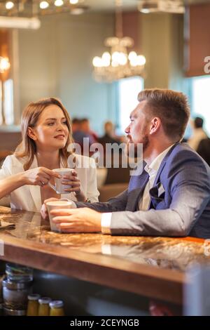 Cheerful long haired brunette woman sits a bar counter, holds a cup of tea, looks with tender smile at impressive man in bright lighted restaurant, cl Stock Photo