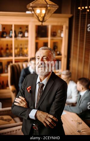 Impressive bearded old man looks happily towards camera with mouth opened, keeps hands crossed over, on background of lighted office Stock Photo