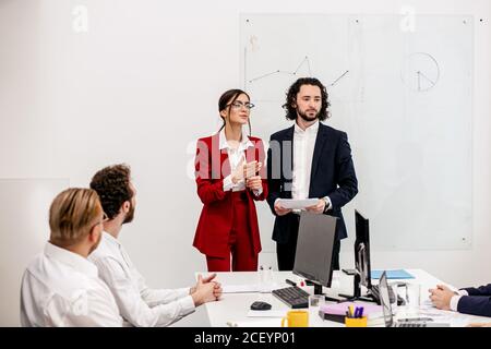 two young caucasian managers together with co-workers in modern office, man in tux and woman in red formal blazer Stock Photo