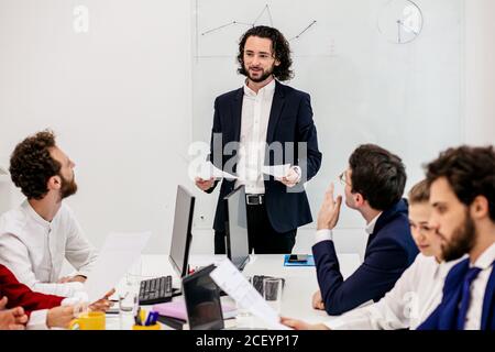 good-looking smiling positive caucasian man manager in tuxedo with co-workers in light office, speak with them holding papers in hands, flipchart in t Stock Photo