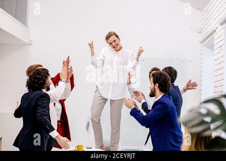 young caucasian business woman dance on table in modern office, celebrating successful completion of her business project, while co-workers support, a Stock Photo