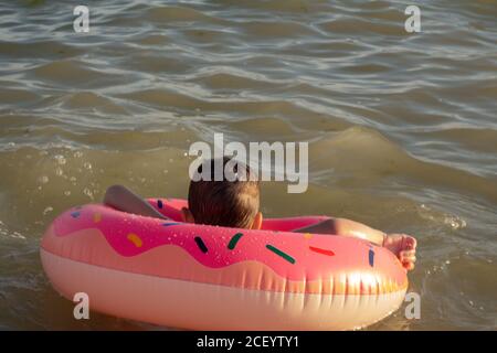 A 5 year old boy swims in the sea, rejoices and has fun in an inflatable ring in the shape of a donut. Stock Photo