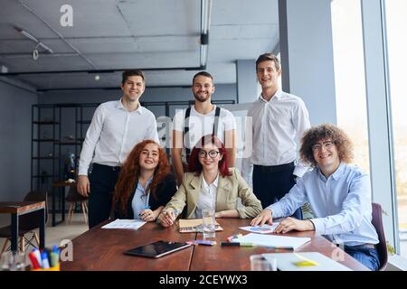 Smiling business team co-working sitting on table together and look at camera, wearing formal wear. Stock Photo