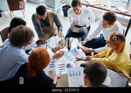 Diverse young people of business team gathered to discuss business startup in modern office, sit together in circle holding papers with diagrams. acti Stock Photo