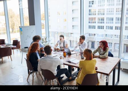Young colleagues, caucasian business partners working as designers sit together at table in modern office with panoramic window. Papers, colorful penc Stock Photo