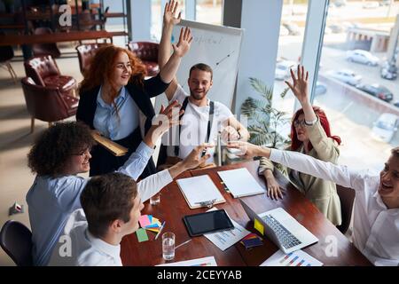 Happy group of colleagues joined hands together finishing up successful business meeting in conference office, papers, colorful pencils on table. Crea Stock Photo