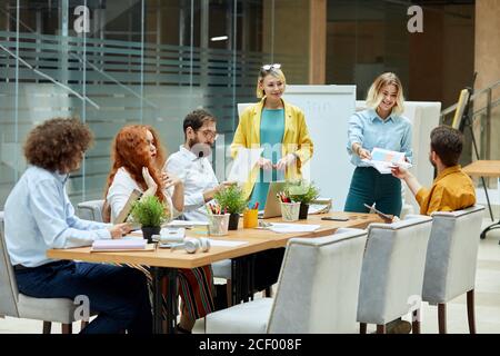Positive, delighted female with wavy hair, dressed casually, stretches documents to handsome bearded young man with broad smile. Creative agency conce Stock Photo
