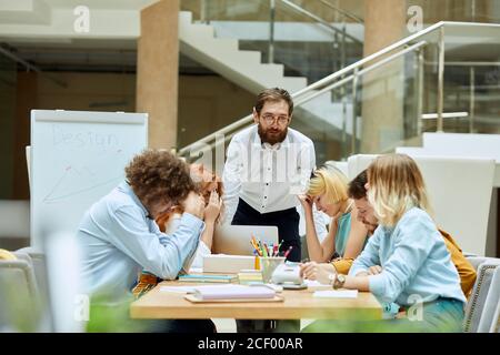 Exacting beard executive with optical spectacles, dressed in white t shirt and dark blue trousers, leans on table with serious face, dissatisfied with Stock Photo