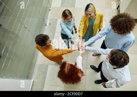 Photo of cheerful young people in bright coloured clothes, standing in brightly lighted office, against glass wall, keeping hands together showing uni Stock Photo