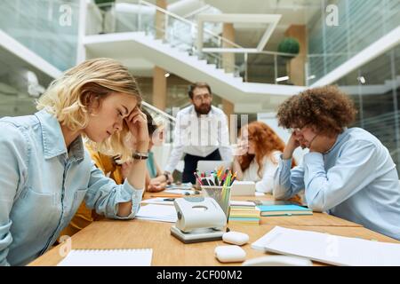 Serious concentrated young female designer, sits at light coloured table, thoughtfully stares at papers, puts elbow on desk , keeps head with delicate Stock Photo