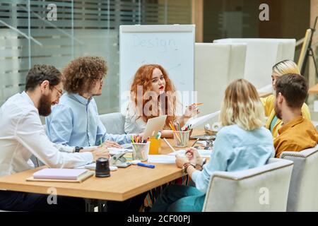 Charming foxy haired woman works together with graphic designers, sits at table, keeps pencil in hand with thoughtful face, team work concept Stock Photo