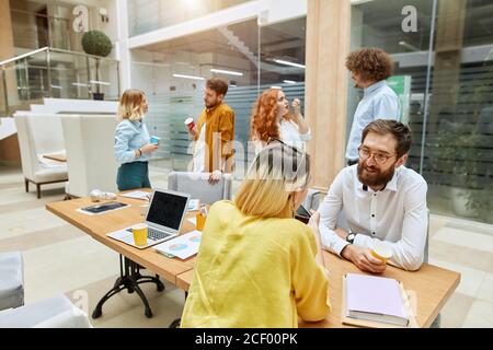 Motivated awesome man with thick beard and optical glasses, wears white long sleeve shirt, sits at working table, holds paper cup, talks to client in Stock Photo