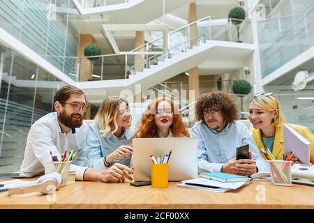 Optimistic young interior designers sit in front of laptop computer, stares at screen with cheerful expression, receive pleasant news from client in m Stock Photo
