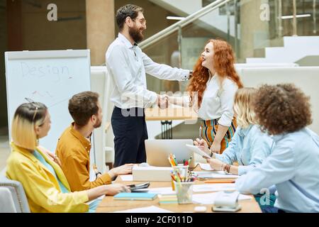 Satisfied powerful team leader in round glasses shake hand with charming touched woman, respectfully puts hand on shoulder at meeting of designers Stock Photo