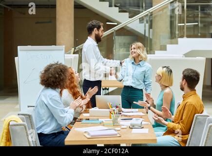 Graphic designers sit in cozy gray chairs at table full of business materials , turn heads towards happy colleagues shaking hands with positive emotio Stock Photo